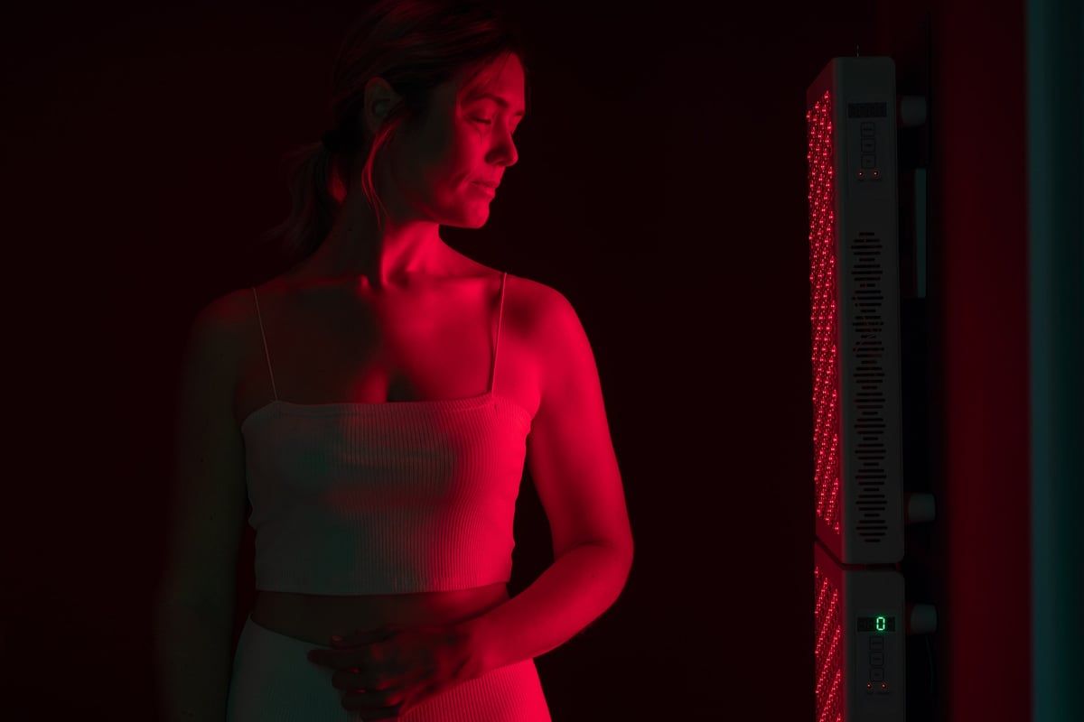 Mature woman receiving red light therapy standing beside a full-body red light panel in a spa setting