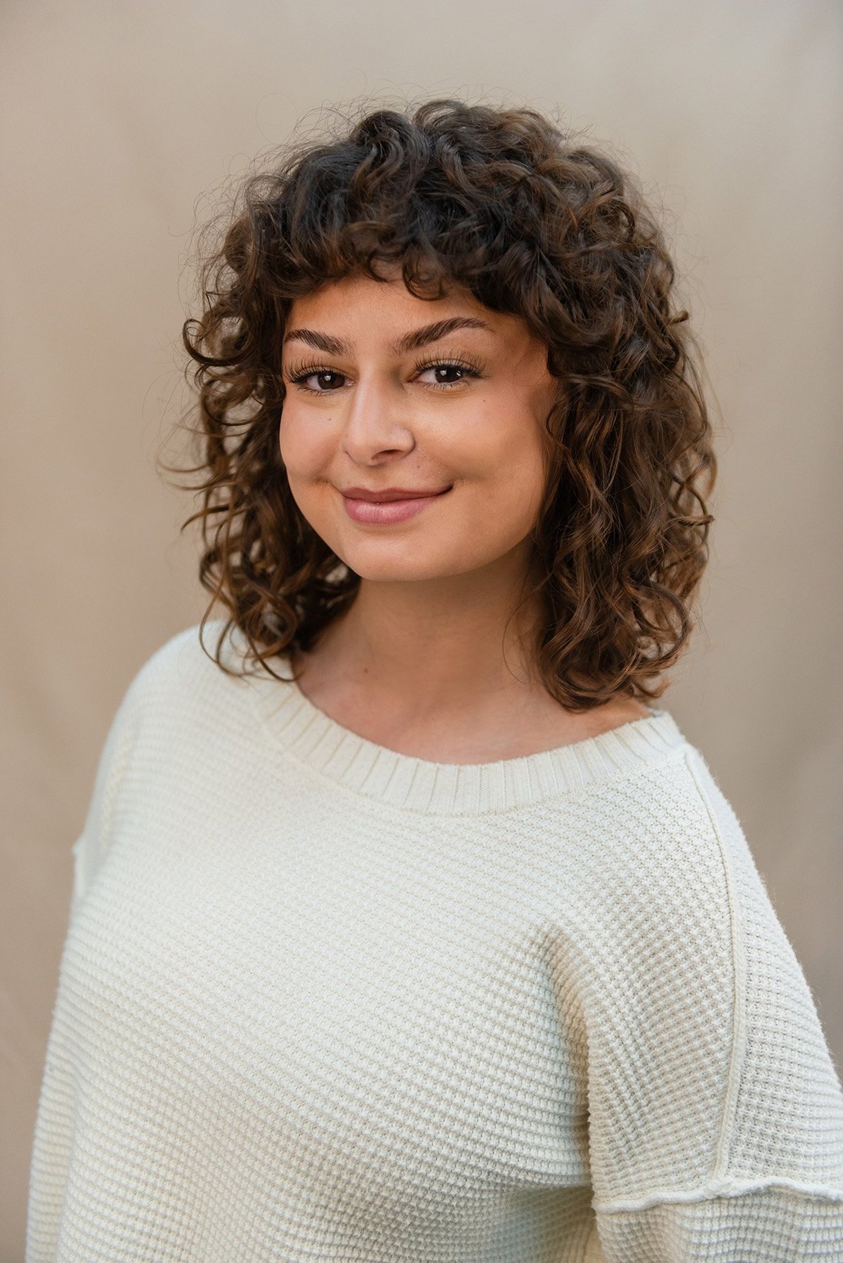 Portrait of Nicole Mendez-Arias, a woman with curly dark brown hair and bangs, wearing a white textured sweater, smiling at the camera