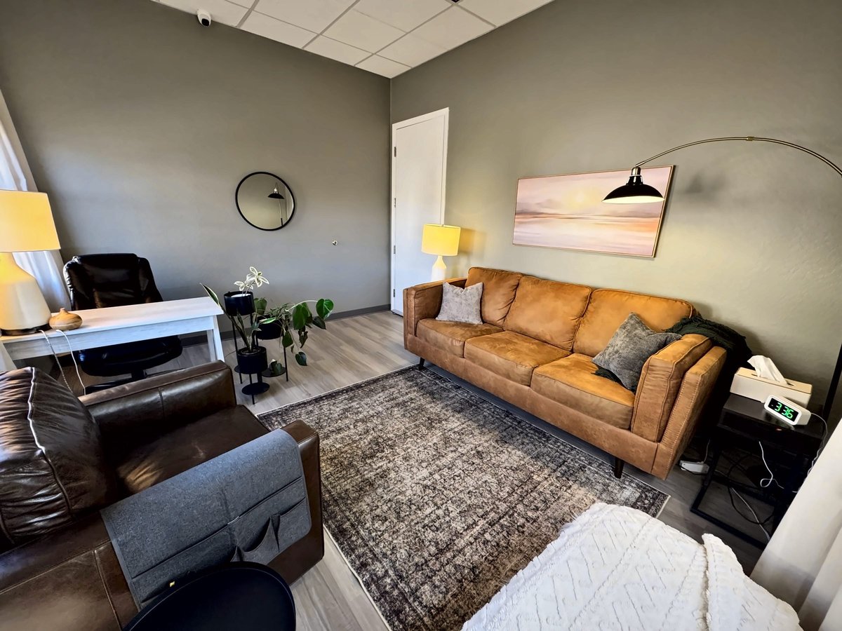 Modern office waiting room with tan leather sofa, black chair, white desk, potted plant, gray area rug, and arc floor lamp.