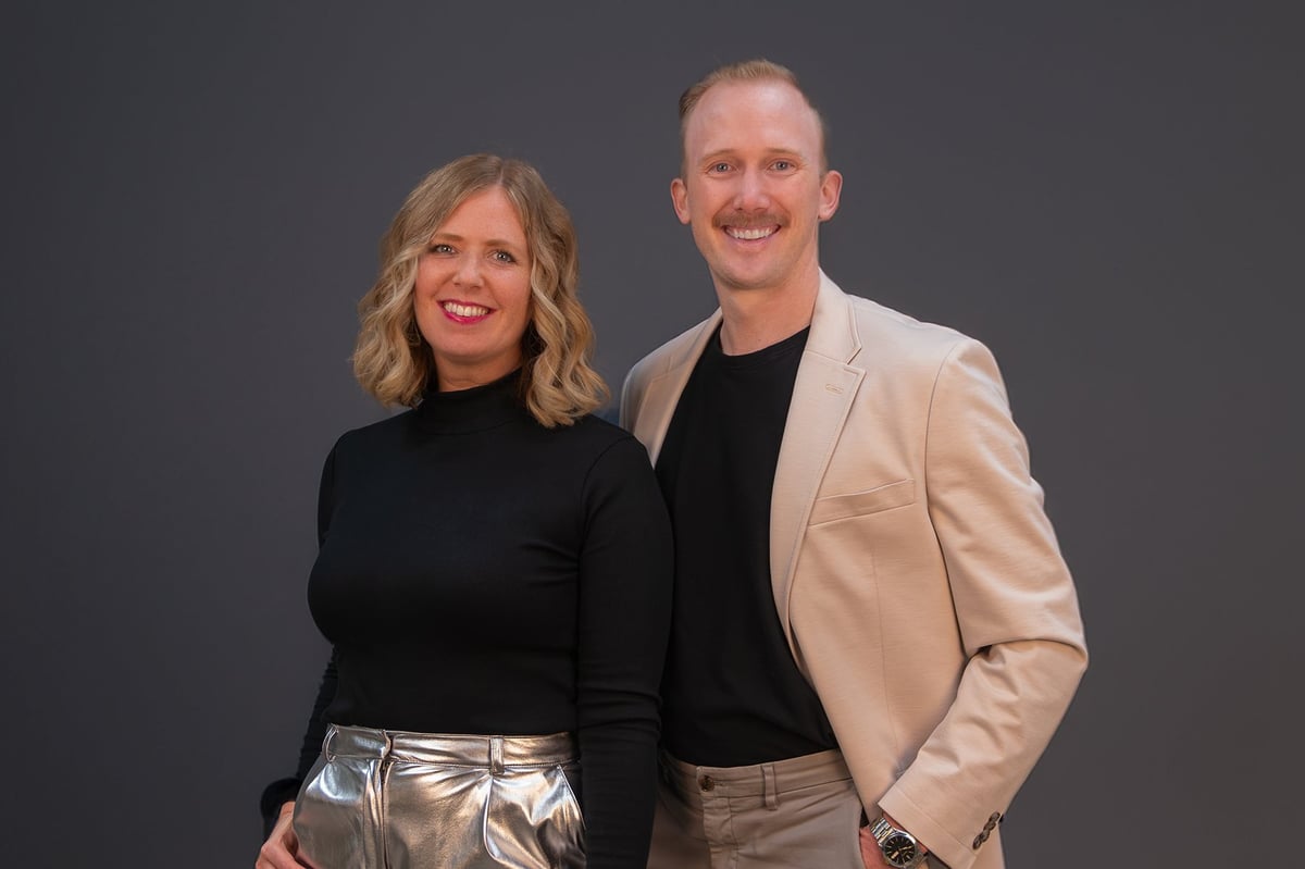 Professional portrait of a woman with blonde wavy hair and red lips in black top with silver pants, and a man in beige blazer and black shirt, both smiling against gray background.