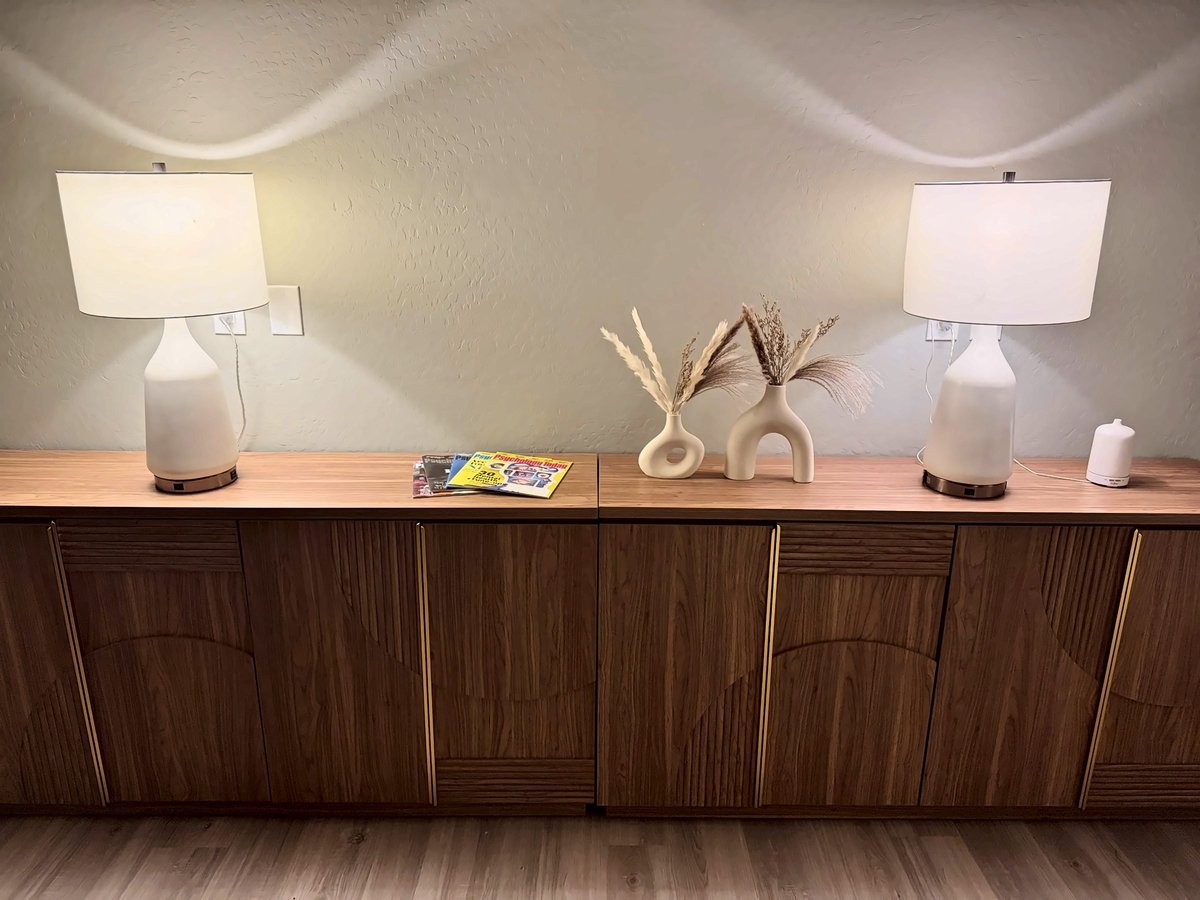 Modern wooden credenza with white table lamps, decorative vases, and magazines in a minimalist room with beige walls