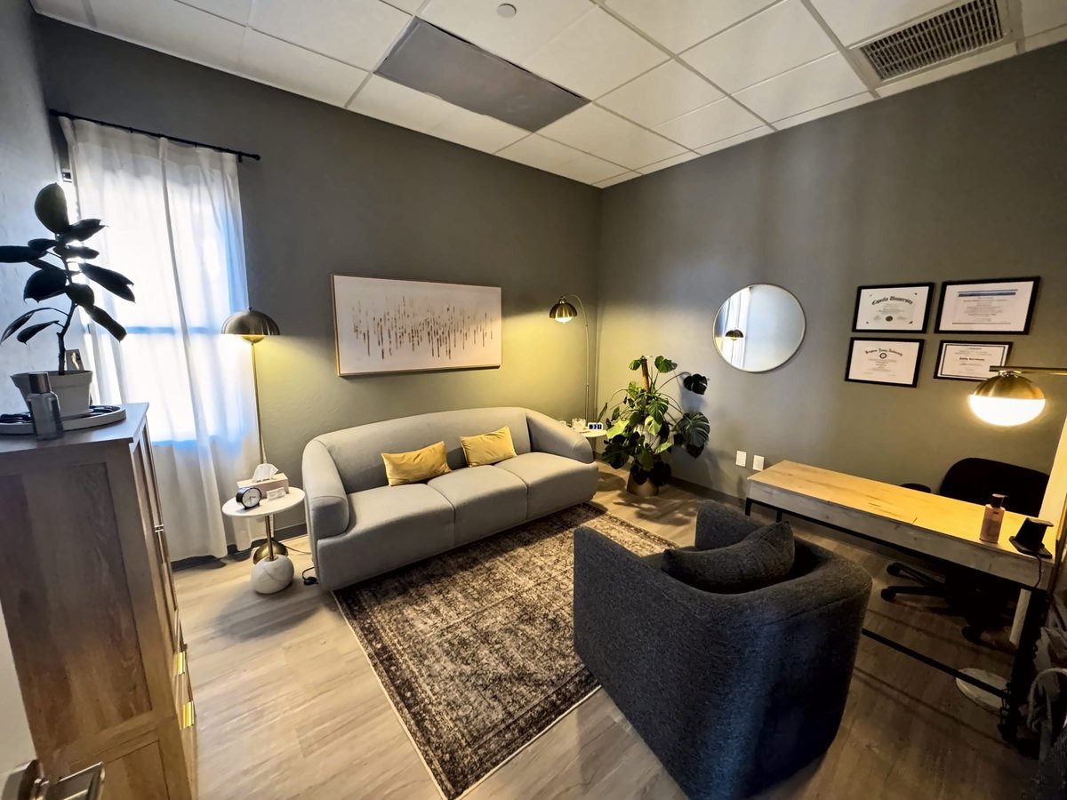Modern office lounge with gray sofa, dark armchair, desk, warm lighting, and potted plants on wooden flooring