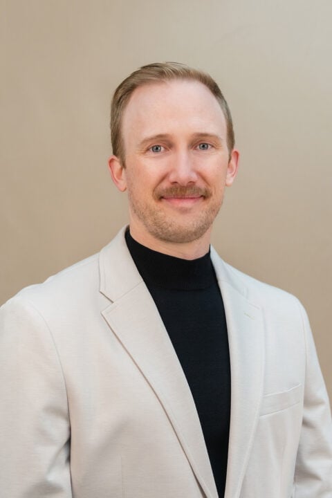 Portrait of Nick Puchalski, a man with blonde hair and mustache wearing a cream blazer over a black turtleneck