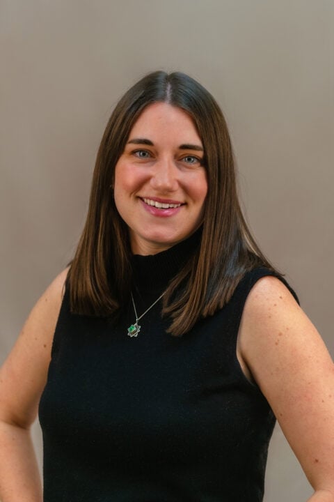 Portrait of Alice Veirs, a smiling woman with long brown hair wearing a black sleeveless top and pendant necklace