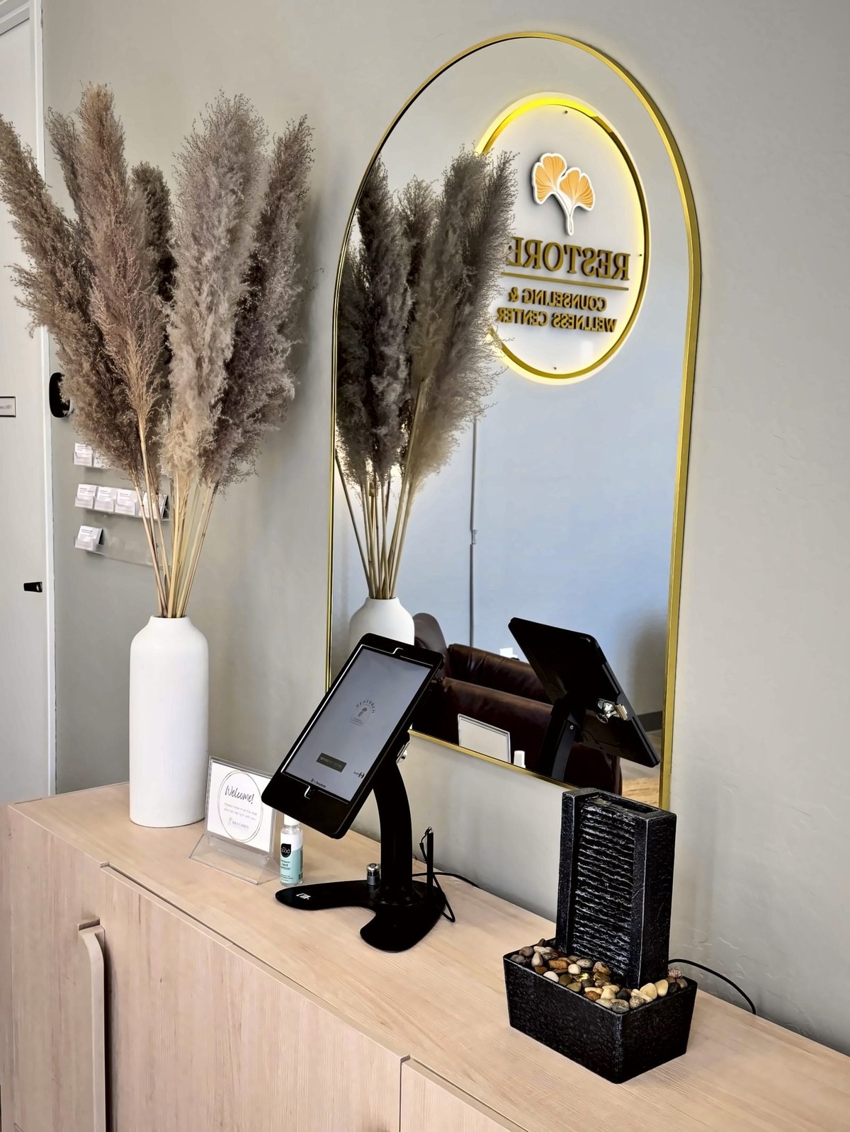 Salon checkout counter with white pampas grass arrangement, gold arch mirror with logo, and black card payment terminal on wooden desk