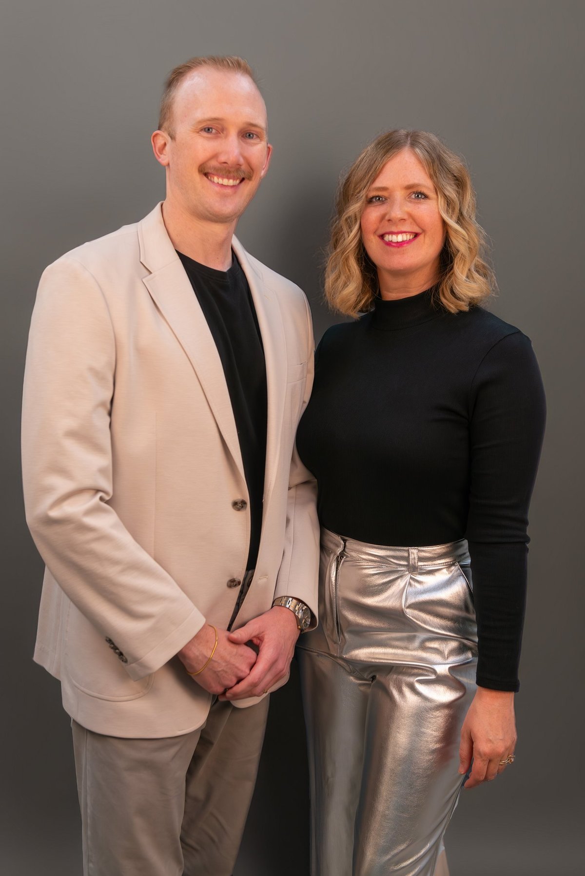 Two people smiling at the camera against a gray background. Man wearing beige blazer and black shirt, woman wearing black top and silver metallic skirt.