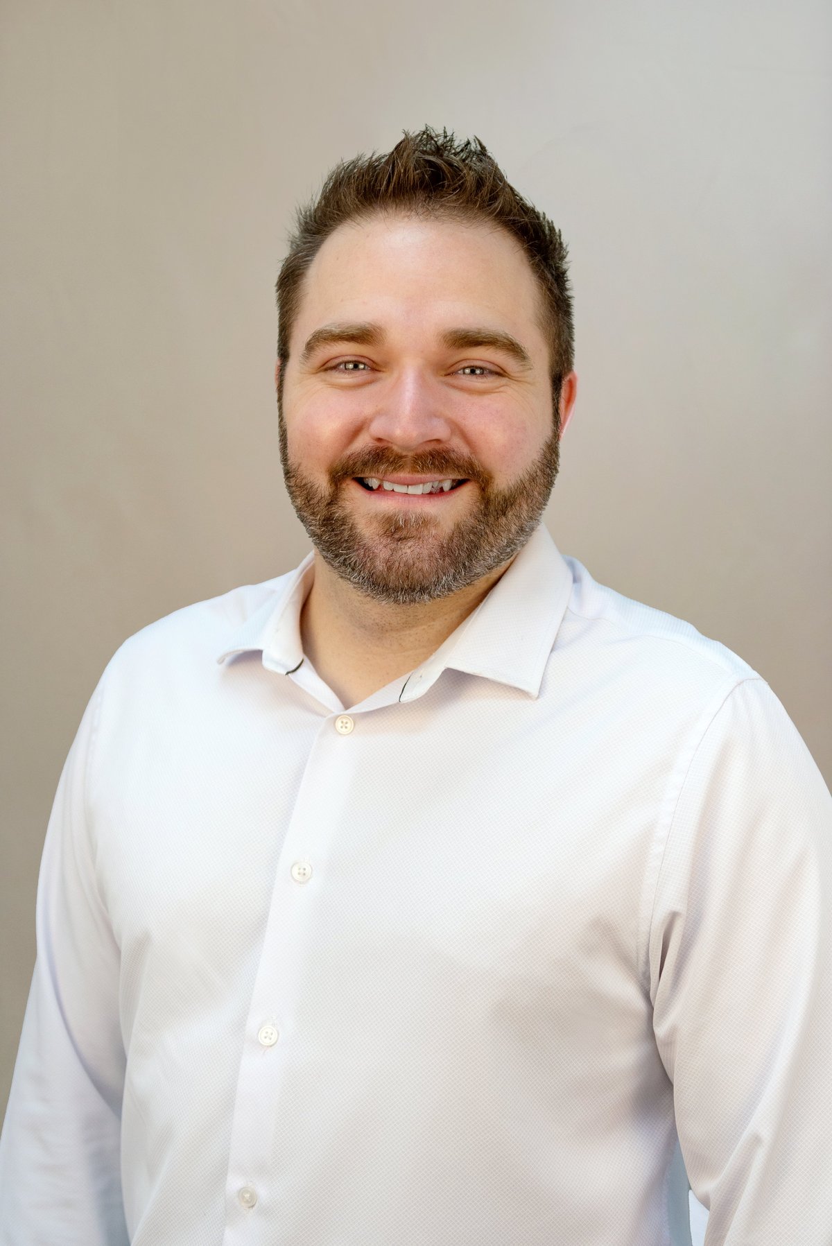 Man with dark hair and beard wearing white button-up shirt, smiling at camera against neutral background