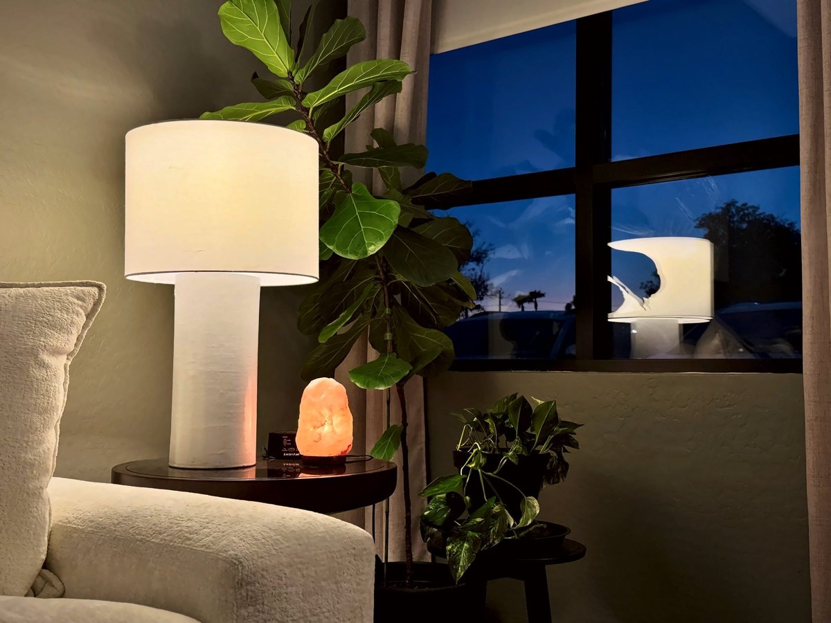 Modern interior with white cylindrical lamp, potted plants, and warm accent lighting beside a window at dusk