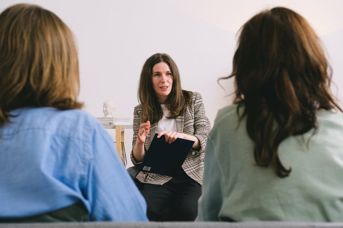 Three women having a conversation in an office setting, with two women facing a woman in the center wearing a checkered jacket